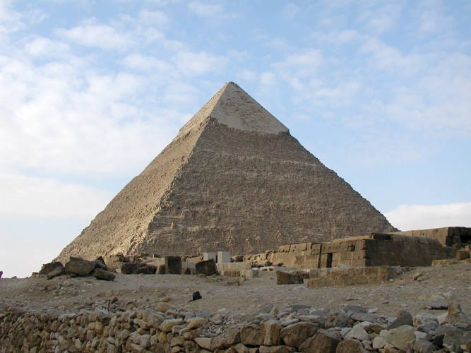 The photo shows the Great Pyramid of Giza in Egypt under a partly cloudy sky, with ancient stone blocks and rubble in the foreground.
