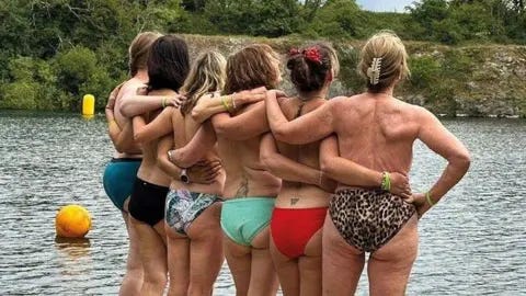 Six women stand on a jetty at Vobster Quay, backs to the camera, wearing bikini bottoms and nothing on top.