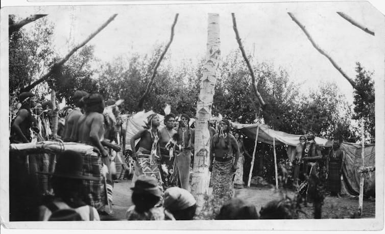 A black-and-white photo showing a ceremony in which several men are standing around a central tree trunk with polelike branches extending outward from it, while a crowd of people are gathered around. A black-and-white photo showing a ceremony in which several men are standing around a central tree trunk with polelike branches extending outward from it, while a crowd of people are gathered around.