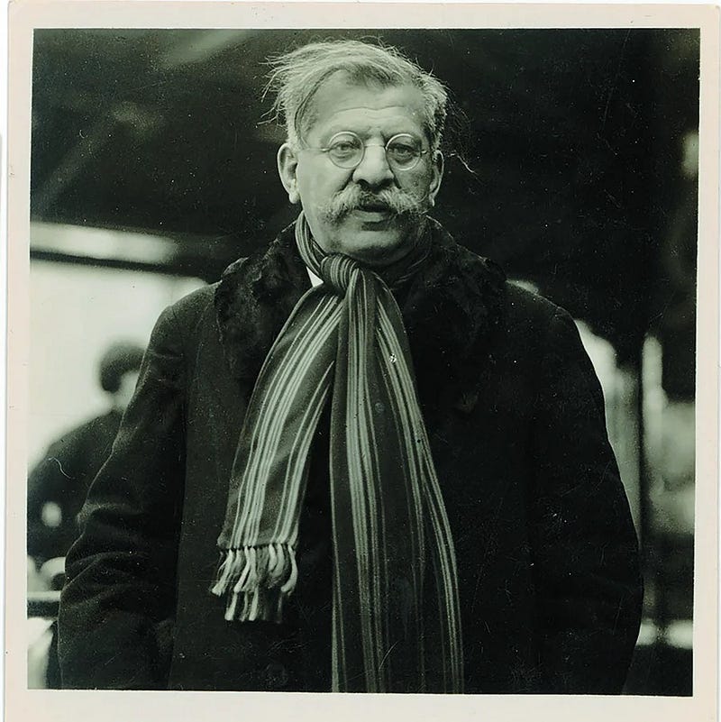 An antique photo of Dr. Magnus Hirschfeld looking at the camera. He is wearing a thick overcoat, a striped scarf, round glasses and a bushy mustache.