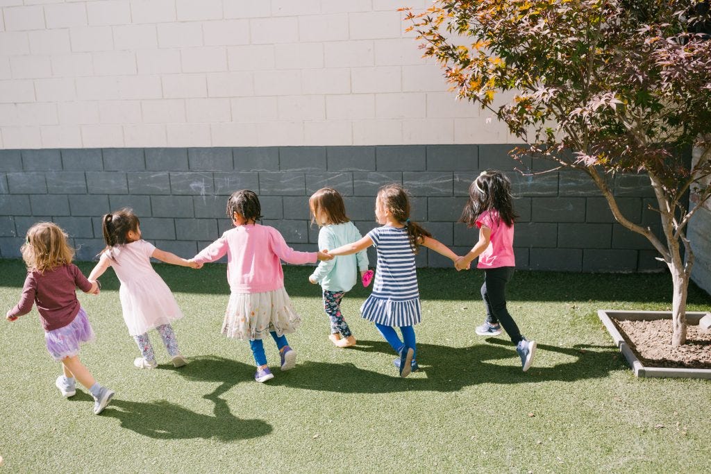 Group of young girls holding hands and running together across a grass playground at a Montessori school