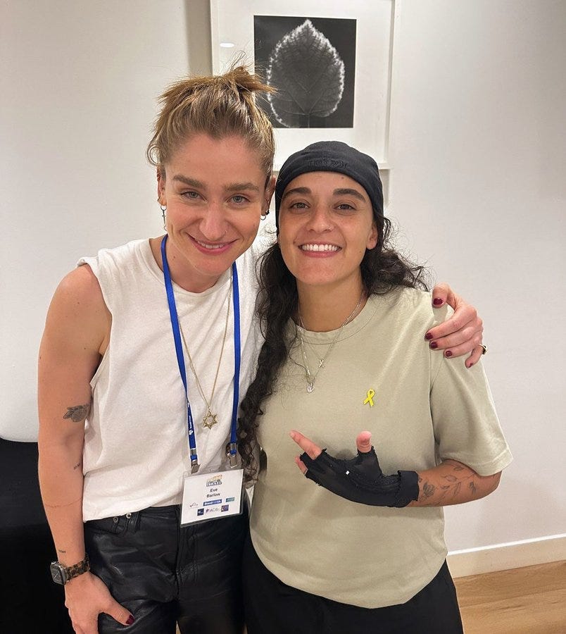 Two women stand side by side smiling at the camera in an indoor setting with white walls and a framed black and white leaf artwork in the background. The left woman has shoulder-length blonde hair tied in a loose bun, fair skin, wears a sleeveless white tank top revealing arm tattoos, a blue lanyard with a name badge around her neck, a necklace, black leather pants, and a wristwatch. The right woman has long wavy dark hair, tan skin, wears a black cap, a loose gray t-shirt with a small yellow ribbon pin on the chest, a necklace, black pants, fingerless black gloves on one hand, a black splint on the other hand showing wrist and finger tattoos, and red nail polish.