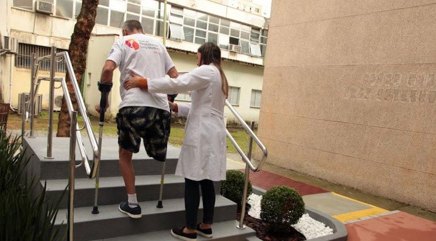 #descrevemospravocê  A imagem mostra um momento de apoio durante uma atividade de reabilitação física. Um homem está subindo uma pequena escada cinza com corrimãos metálicos dos dois lados. Ele usa muletas e tem uma das pernas amputada, usando uma prótese na perna direita. Ao lado dele, uma profissional de saúde, vestindo jaleco branco e calça escura, o acompanha de perto, com a mão nas costas dele, oferecendo suporte.  O cenário é externo, em frente a um prédio de paredes claras com janelas grandes. Ao fundo, há vegetação com grama, uma árvore e uma área com arbustos podados. À direita da imagem, parte da calçada conta com piso tátil amarelo, indicando acessibilidade para pessoas com deficiência visual. O chão tem faixas coloridas em vermelho e bege, e há um pequeno jardim com pedras brancas e arbustos arredondados. Na parede à direita, é possível ver um letreiro em relevo, mas as palavras estão parcialmente desfocadas.