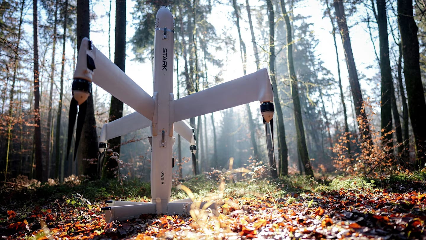 The "Virtus" drone weapon from Stark Defense stands on the forest floor in a wooded area, illuminated by sunlight filtering through trees.