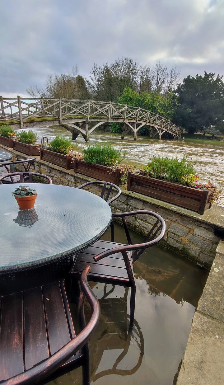 A pub garden terrace flooded with water with the river and a small bridge in the background; a vast expanse of water dotted with islands reflecting a cloudy sky