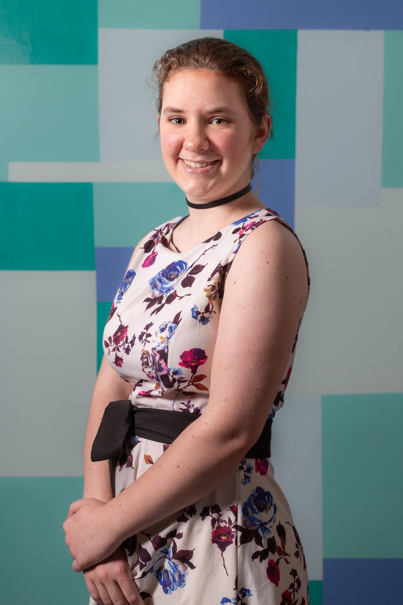 Portrait of a 17 year old, white skinned woman. She has brown hair pulled back and light brown eyes. She is smiling at the camera showing her teeth. She is wearing a black thin choker necklace and a sleeveless floral dress with a black belt.