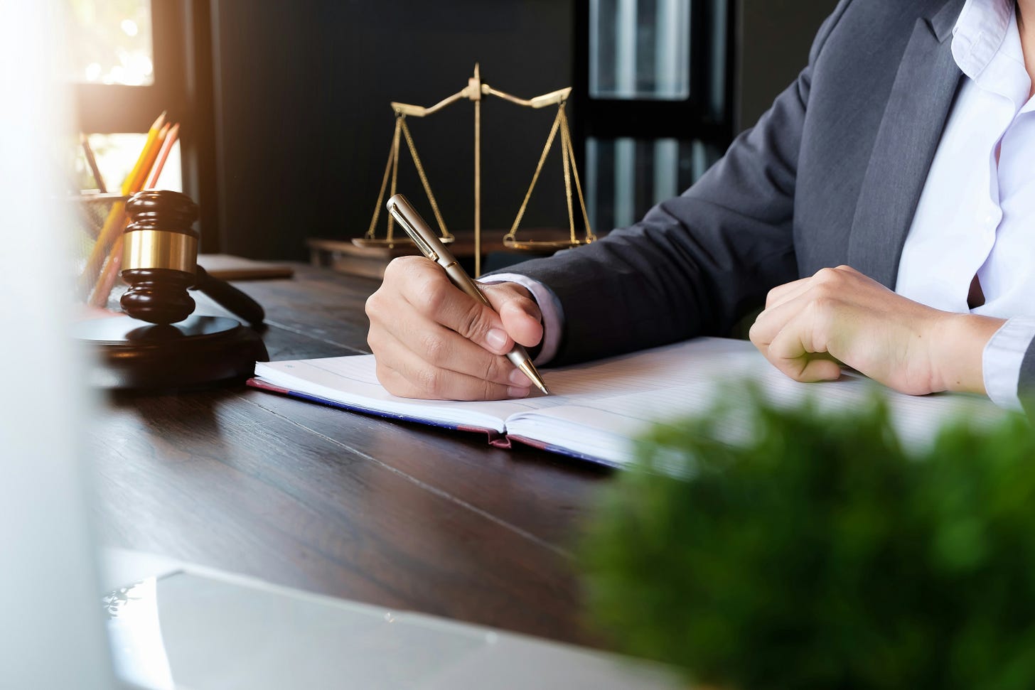 a stock photo of a person at a table writing. they are surrounded by objects evoking ideas of the courts and justice, such as a gavel and scales.