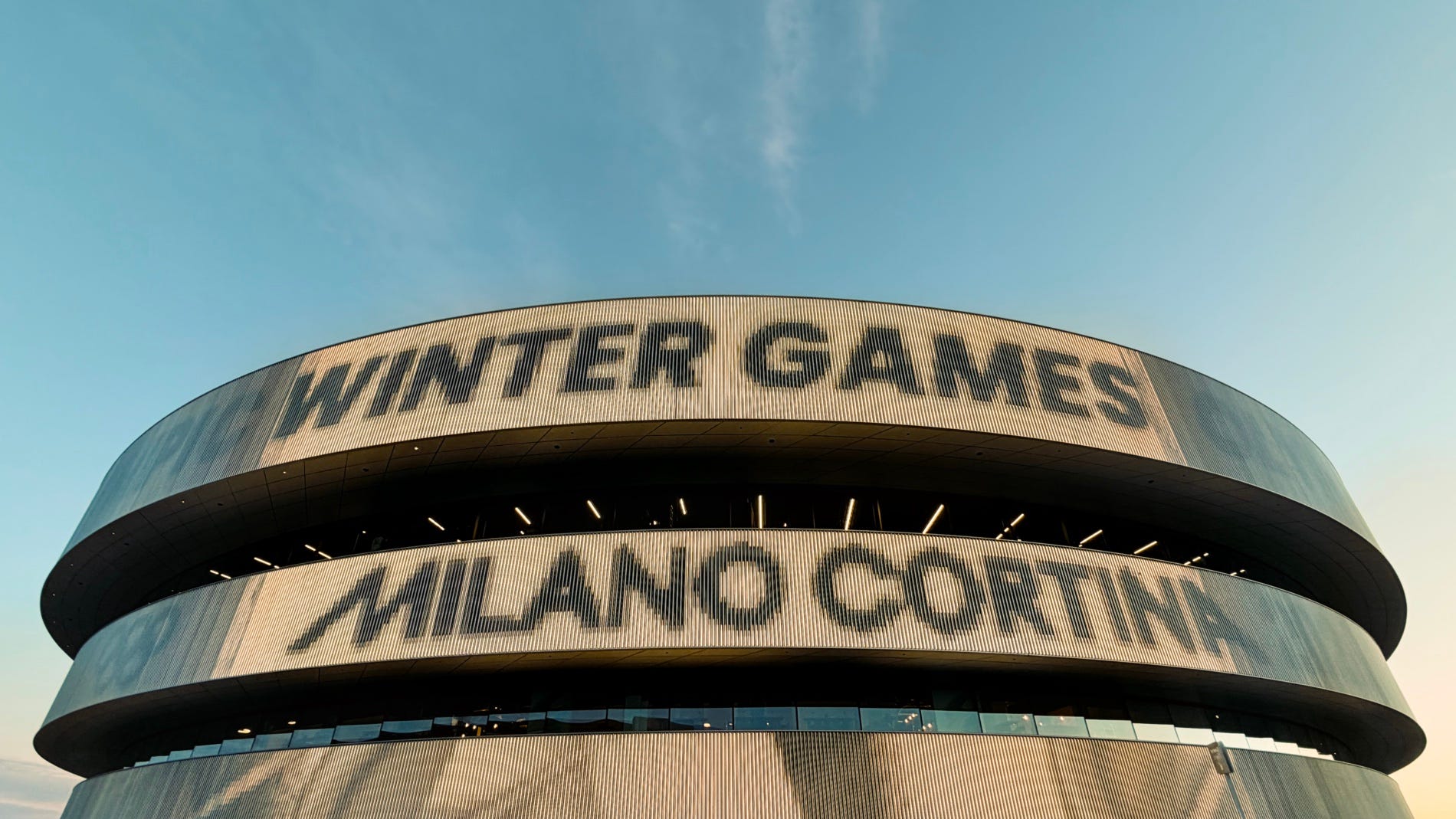 A modern circular arena photographed from below against a clear blue sky, with large bold text reading "WINTER GAMES" and "MILANO CORTINA" displayed across its curved exterior facade, lit by warm evening light. By Swiss and Chps