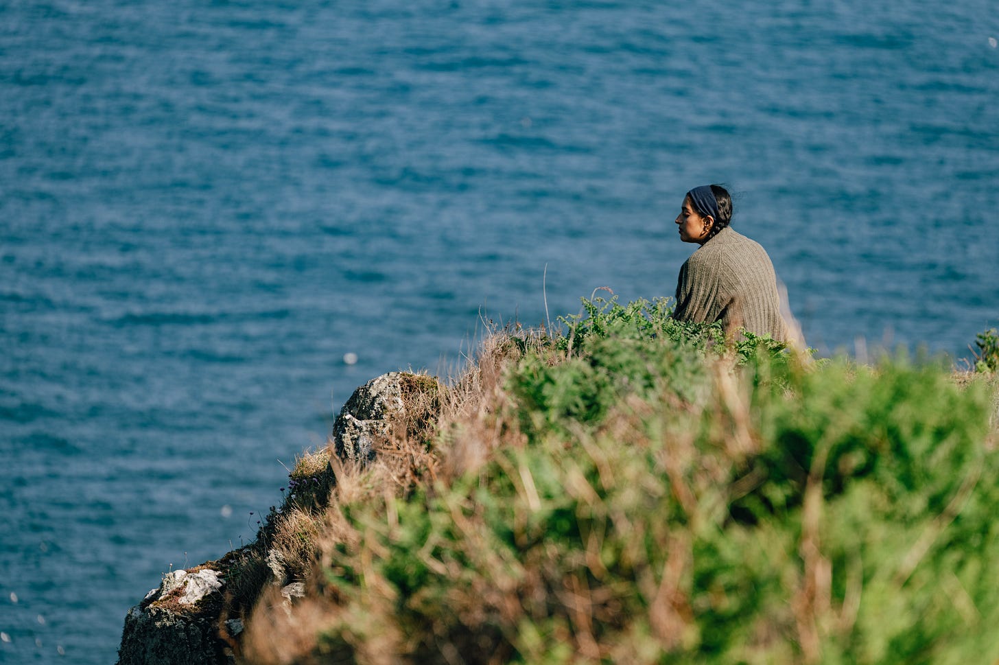 A woman sits near the cliff edge, looking out over the calm blue water, framed by green coastal foliage. A woman sits near the cliff edge, looking out over the calm blue water, framed by green coastal foliage.