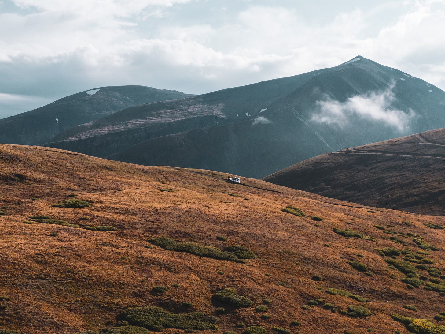 An aerial drone shot of the dark-colored Ford Escape parked on a dirt road, centered in the frame and surrounded by vast mountains.