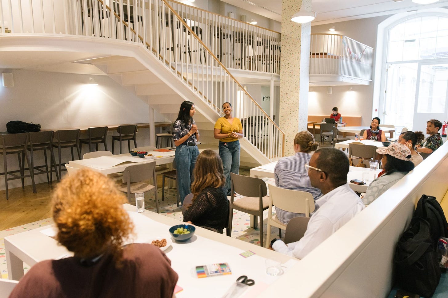 A group of about ten people seated in an open studio looking towards two women standing at the front addressing the group. A group of about ten people seated in an open studio looking towards two women standing at the front addressing the group.