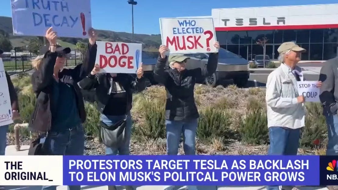 People holding signs reading 'BAD DOGE!' 'WHO ELECTED MUSK?' and 'STOP THE COUP.' Behind them are landscaped desert plants and the car dealership, with two Cybertrucks and a Model Y(?) in the otherwise empty lot. NBC News chyron reads 'Protesters target Tesla as backlash to Elon Musk's political power grows.' One sign is only partly visible; it reads 'Truth Decay,' so it might have been 'Musk (or Trump or Tesla) causes ...' People holding signs reading 'BAD DOGE!' 'WHO ELECTED MUSK?' and 'STOP THE COUP.' Behind them are landscaped desert plants and the car dealership, with two Cybertrucks and a Model Y(?) in the otherwise empty lot. NBC News chyron reads 'Protesters target Tesla as backlash to Elon Musk's political power grows.' One sign is only partly visible; it reads 'Truth Decay,' so it might have been 'Musk (or Trump or Tesla) causes ...'