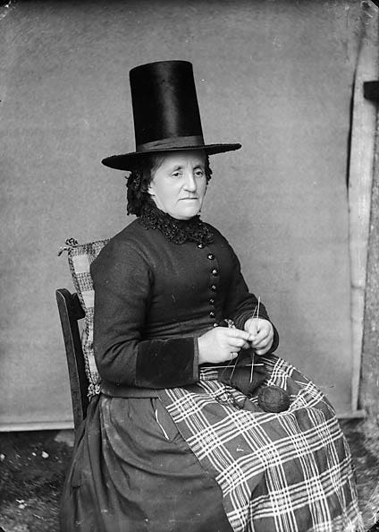 Black and white photograph of a woman in a tall black hat seated in a chair, knitting, with a ball of yarn in her lap.