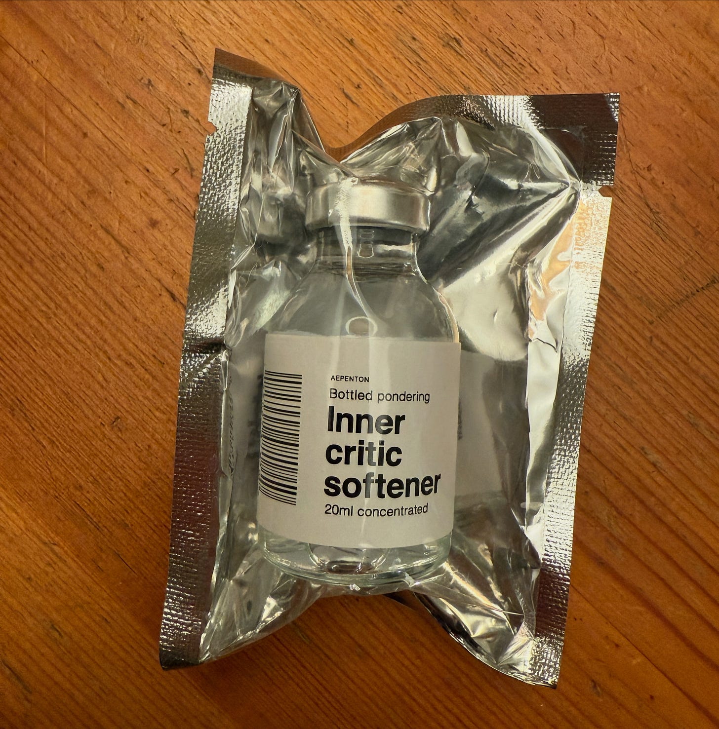 A 20ml vial labeled 'Inner Critic Softener,' part of the Bottled Pondering exhibit by Aepenton at the Design Museum Denmark, sealed in reflective foil packaging. A 20ml vial labeled 'Inner Critic Softener,' part of the Bottled Pondering exhibit by Aepenton at the Design Museum Denmark, sealed in reflective foil packaging.