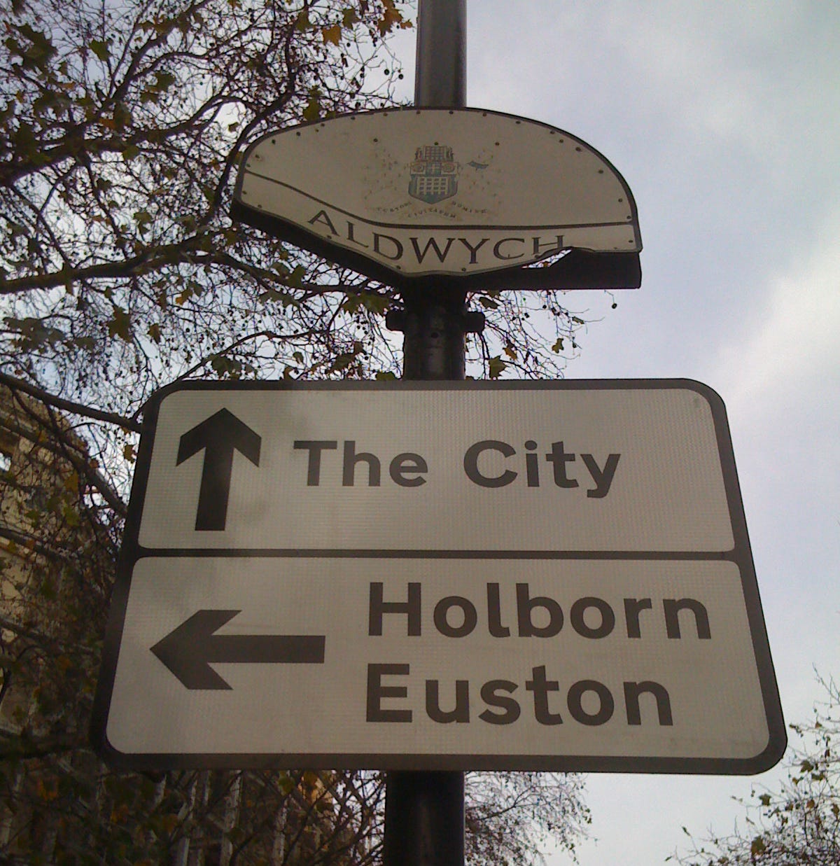Photograph of a modern road sign pointing to The City, Holborn and Euston, mounted below a much older and badly dented semicircular panel that reads ALDWYCH Photograph of a modern road sign pointing to The City, Holborn and Euston, mounted below a much older and badly dented semicircular panel that reads ALDWYCH