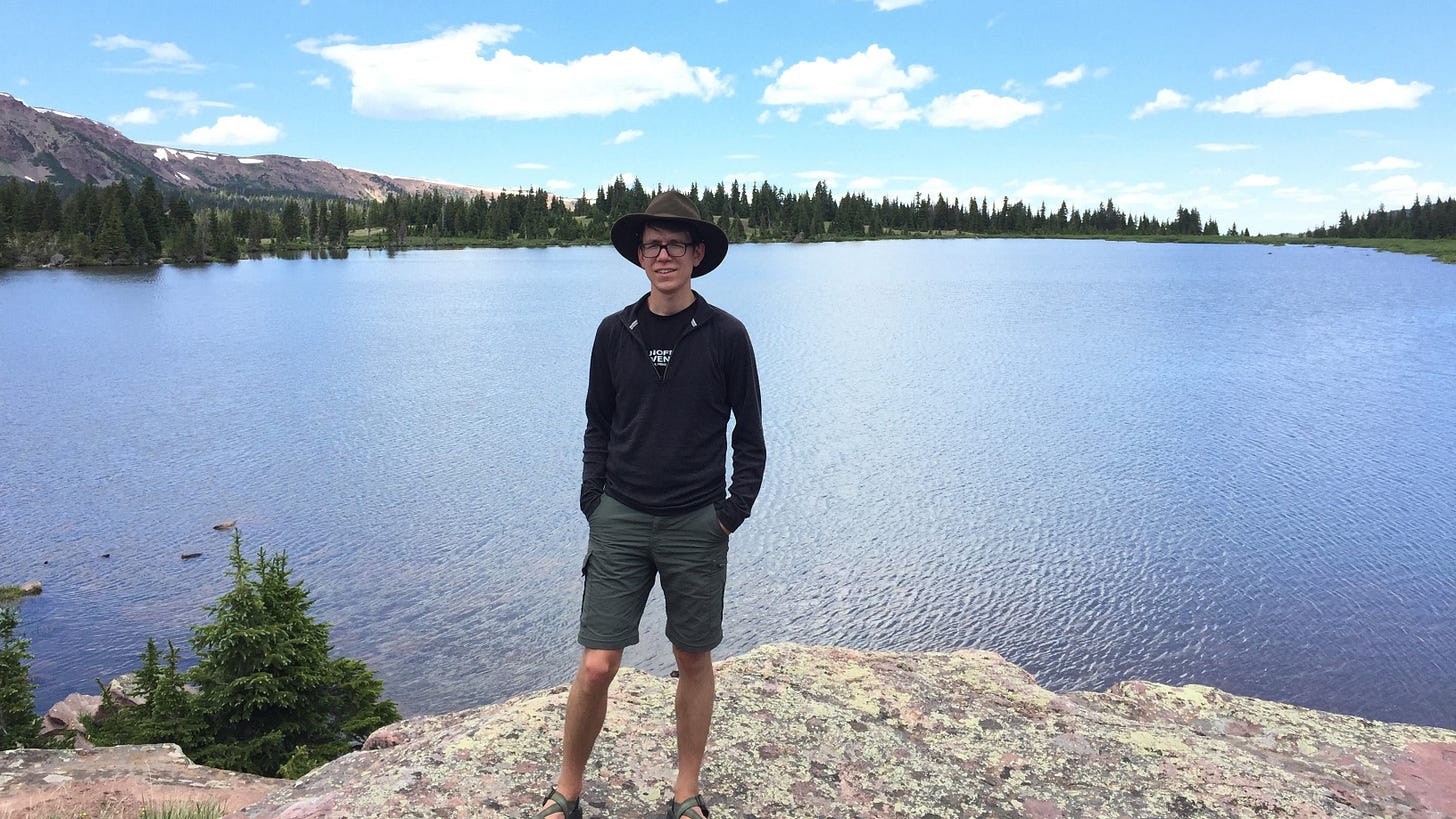 A man wearing a felt hat stands in front of a mountain lake.