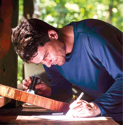 Pessoa usando camiseta azul marinho inclinada sobre uma superfície de madeira clara, segurando um instrumento de trabalho na mão direita enquanto escreve ou marca algo na madeira com a mão esquerda, com fundo desfocado de vegetação verde e iluminação natural.