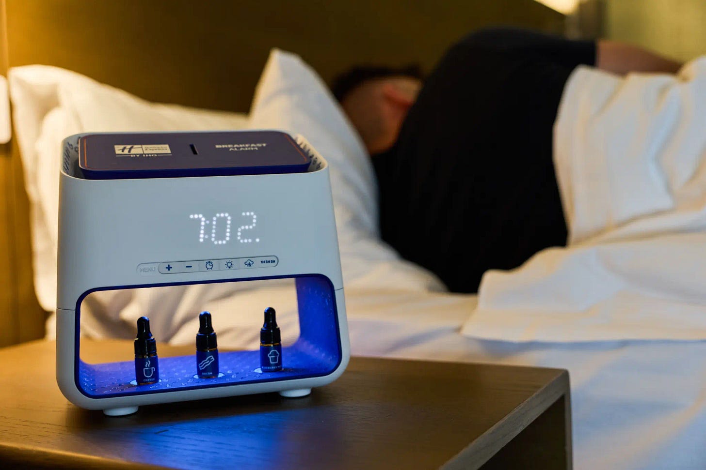 A person asleep in a hotel bed next to a nightstand with an alarm clock; the alarm clock is open at the bottom and three tiny bottles with eyedroppers sit inside. Each has an icon on the front, coffee, bacon, and muffin