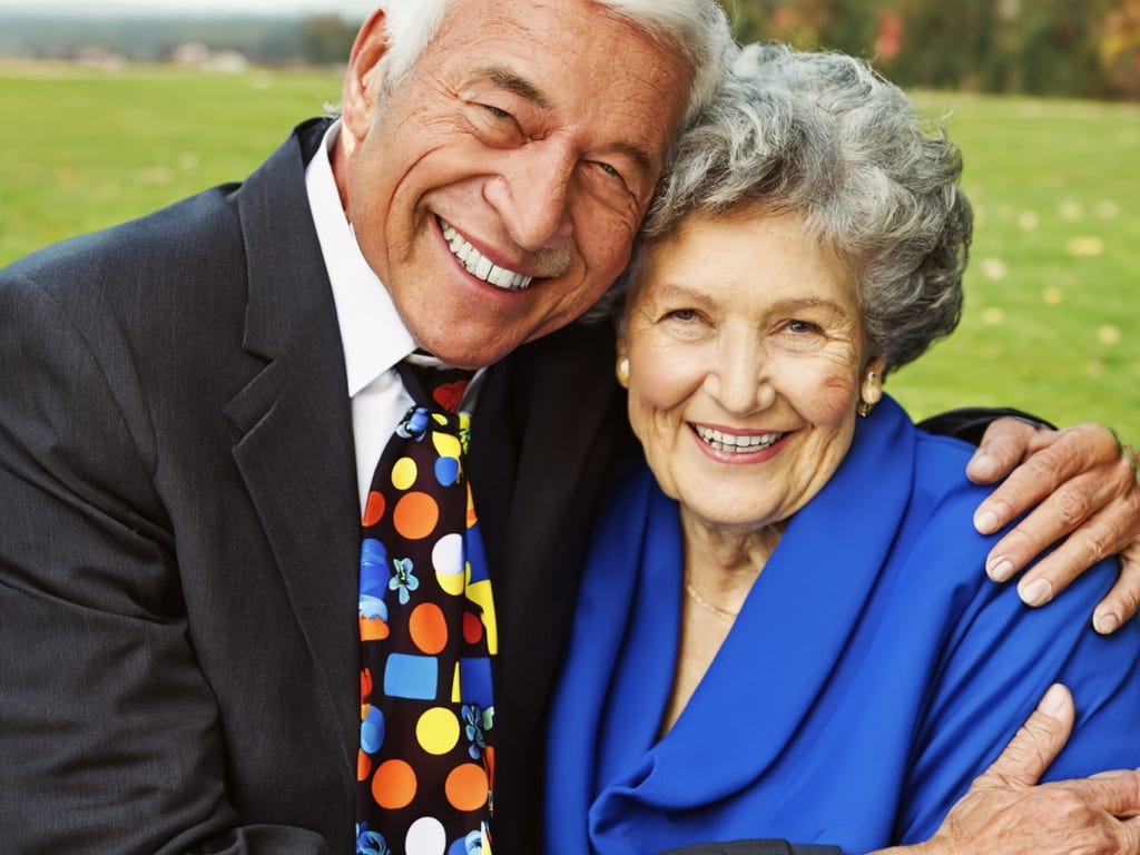 80-year-old, gray-haired couple in embrace, smiling, he in suit with polka-dotted tie and she in blue gown.