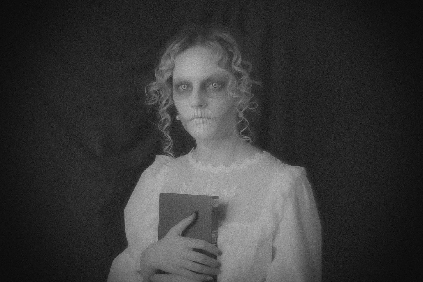 A monochrome photo of a pale woman wearing a white dress and holding a book, staring out at the viewer. She is wearing skeleton-like facial makeup, giving her face an eerie counternance. Image credit: copyright Ksenia Yakovleva from Unsplash.