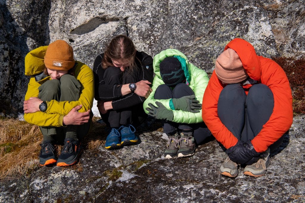 Four people crouch in a row against a rock face, knees drawn up and faces tucked down, lit by low warm evening light.