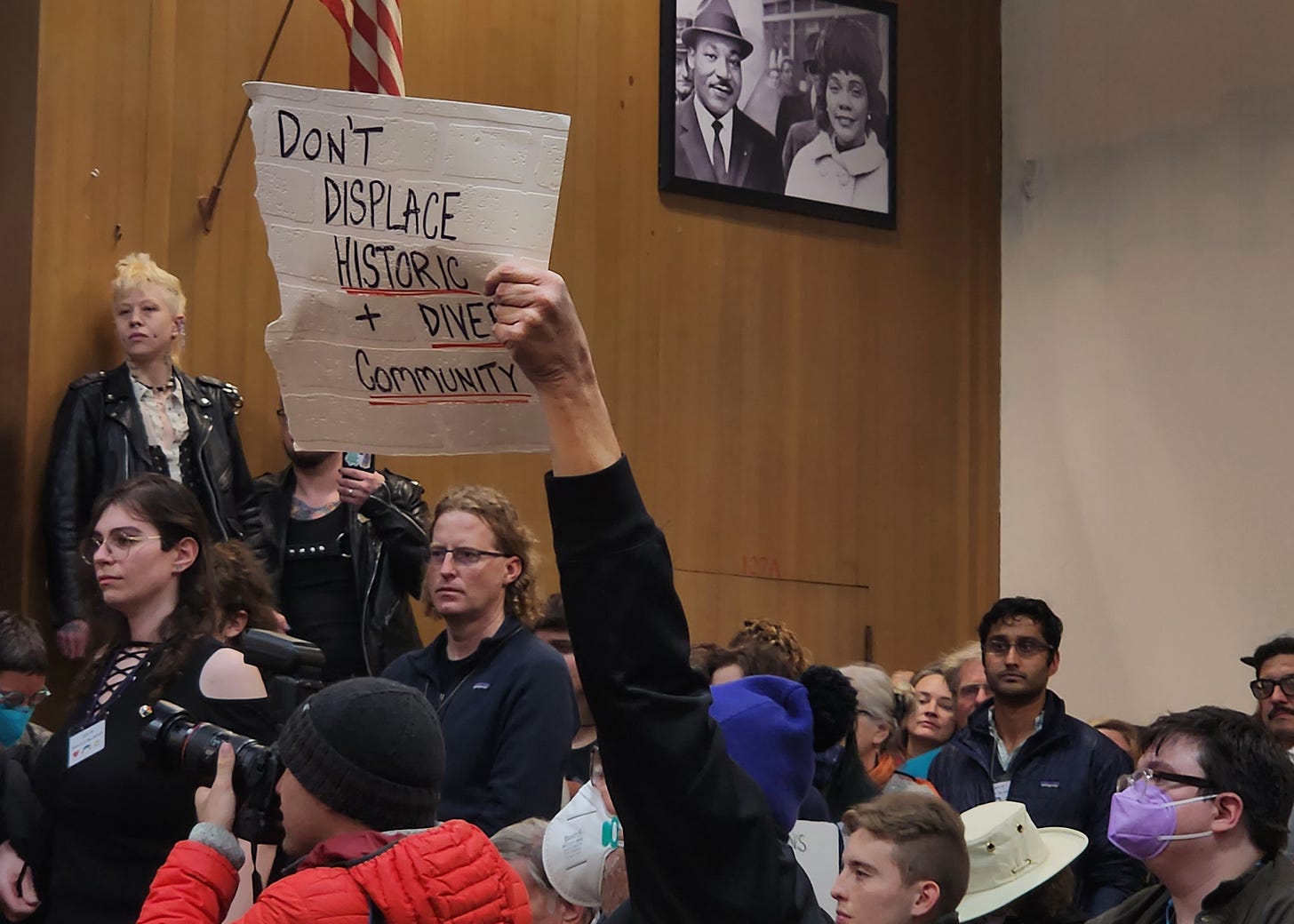 A packed community meeting room where a person holds up a handwritten cardboard sign reading "Don't displace historic + diverse community." A crowd of attendees fills the room behind them, with a photograph of Martin Luther King Jr. visible on the wood-paneled wall. A person in a black leather jacket stands on a chair in the background. A photographer with a camera documents the scene in the foreground. A packed community meeting room where a person holds up a handwritten cardboard sign reading "Don't displace historic + diverse community." A crowd of attendees fills the room behind them, with a photograph of Martin Luther King Jr. visible on the wood-paneled wall. A person in a black leather jacket stands on a chair in the background. A photographer with a camera documents the scene in the foreground.