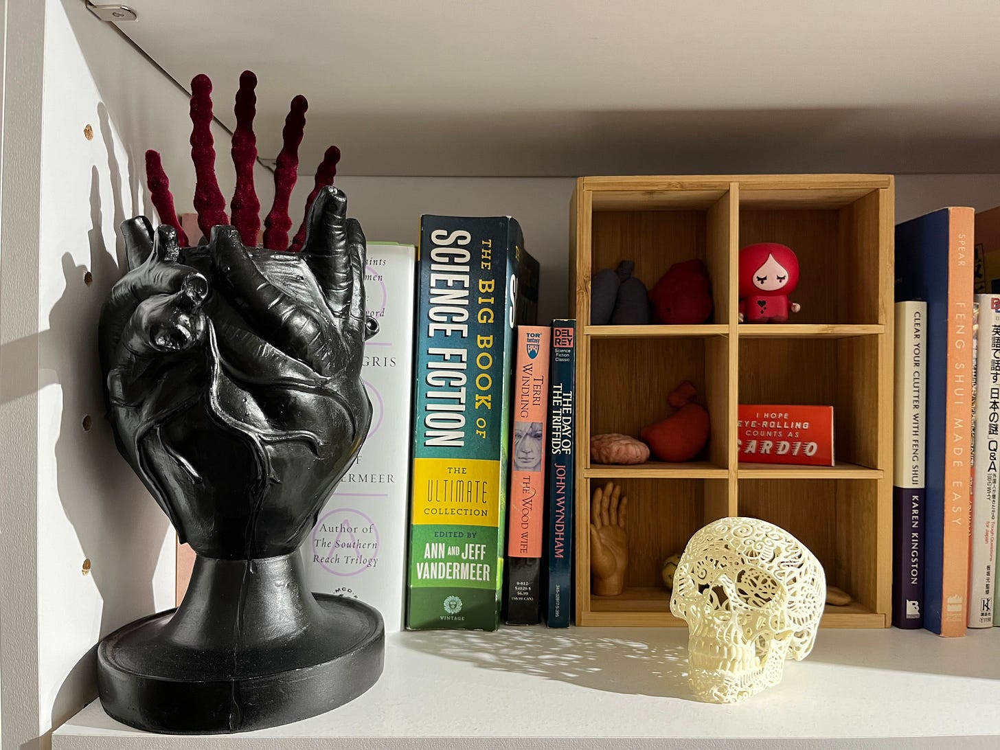 One of Lia's studio shelves. A black plaster anatomical heart vase sits on the left, with a burgundy velvet skeletal hand sticking out of it. There's also a delicate filigree skull on the shelf in front of a tiny curio box with various small items in it. Some science fiction books are also visible. 