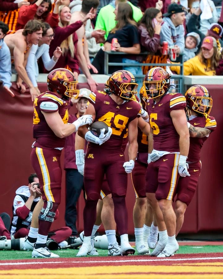 Matt Kingsbury wearing a maroon and gold football uniform with the number 49, celebrating on the field with three other players in matching uniforms. They are holding a football, with a crowd visible in the background. The players are wearing helmets and white gloves. Matt Kingsbury wearing a maroon and gold football uniform with the number 49, celebrating on the field with three other players in matching uniforms. They are holding a football, with a crowd visible in the background. The players are wearing helmets and white gloves.