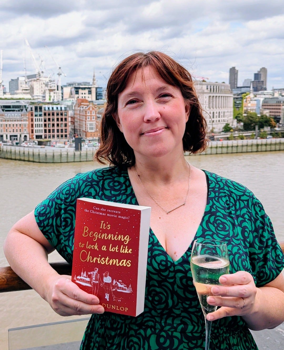 Hayley stands on a Southbank roof terrace holding a copy of her book and a glass of prosecco