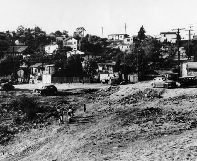 People climb a dirt hill in the foreground toward houses on the hillside.