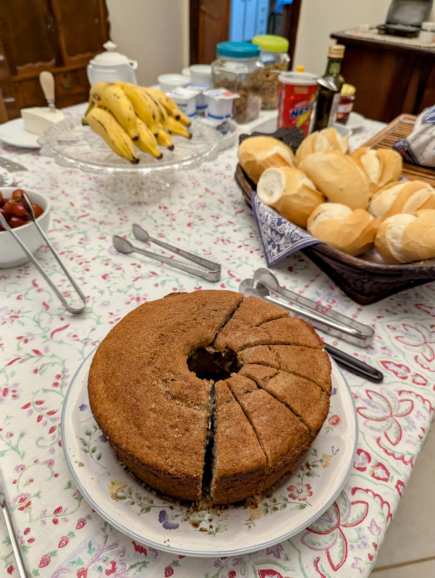 A breakfast spread, showing a sliced cake, breads, bananas, and utensils to pick up items. A breakfast spread, showing a sliced cake, breads, bananas, and utensils to pick up items.