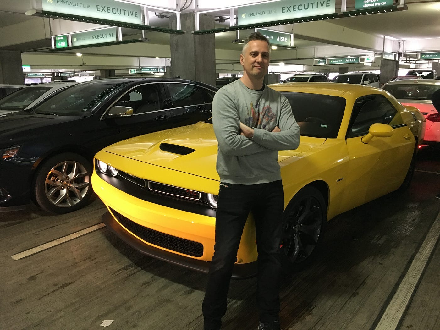 A man standing arms folded in front of a bright yellow Dodge Challenger muscle car in a National car rental car park, the Emerald Club Executive signs visible overhead. The expression says this was entirely intentional. A man standing arms folded in front of a bright yellow Dodge Challenger muscle car in a National car rental car park, the Emerald Club Executive signs visible overhead. The expression says this was entirely intentional.