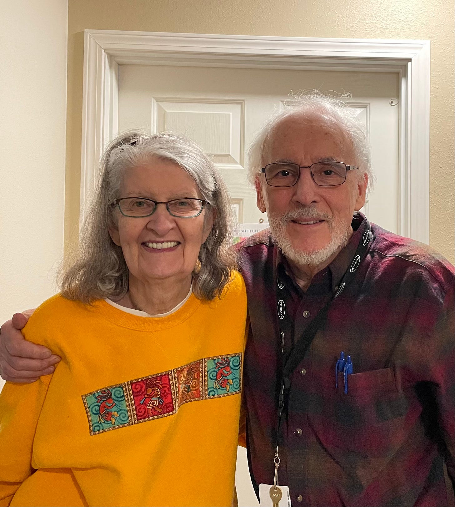 And older couple, arm in arm, smiling in front of a white door 
