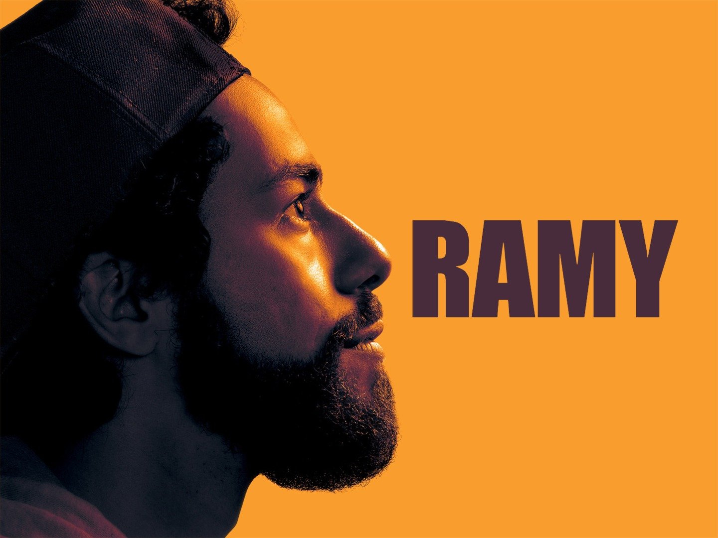 La locandina di Ramy con il primo piano dell'attore Ramy Youssef, ragazzo di origine egiziana con la barba scura e un cappellino da baseball al contrario. È fotografato di profilo e il suo viso è illuminato da una calda luce gialla, simile allo sfondo della locandina.