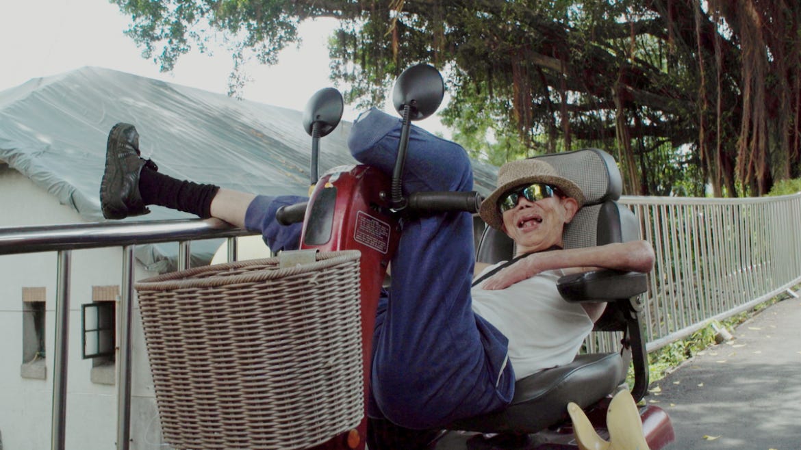 An elderly man dressed in a white tank top, blue sweatpants, sunglasses, and a straw hat, reclines in the seat of a mobility scooter. His left leg is amputated below the knee