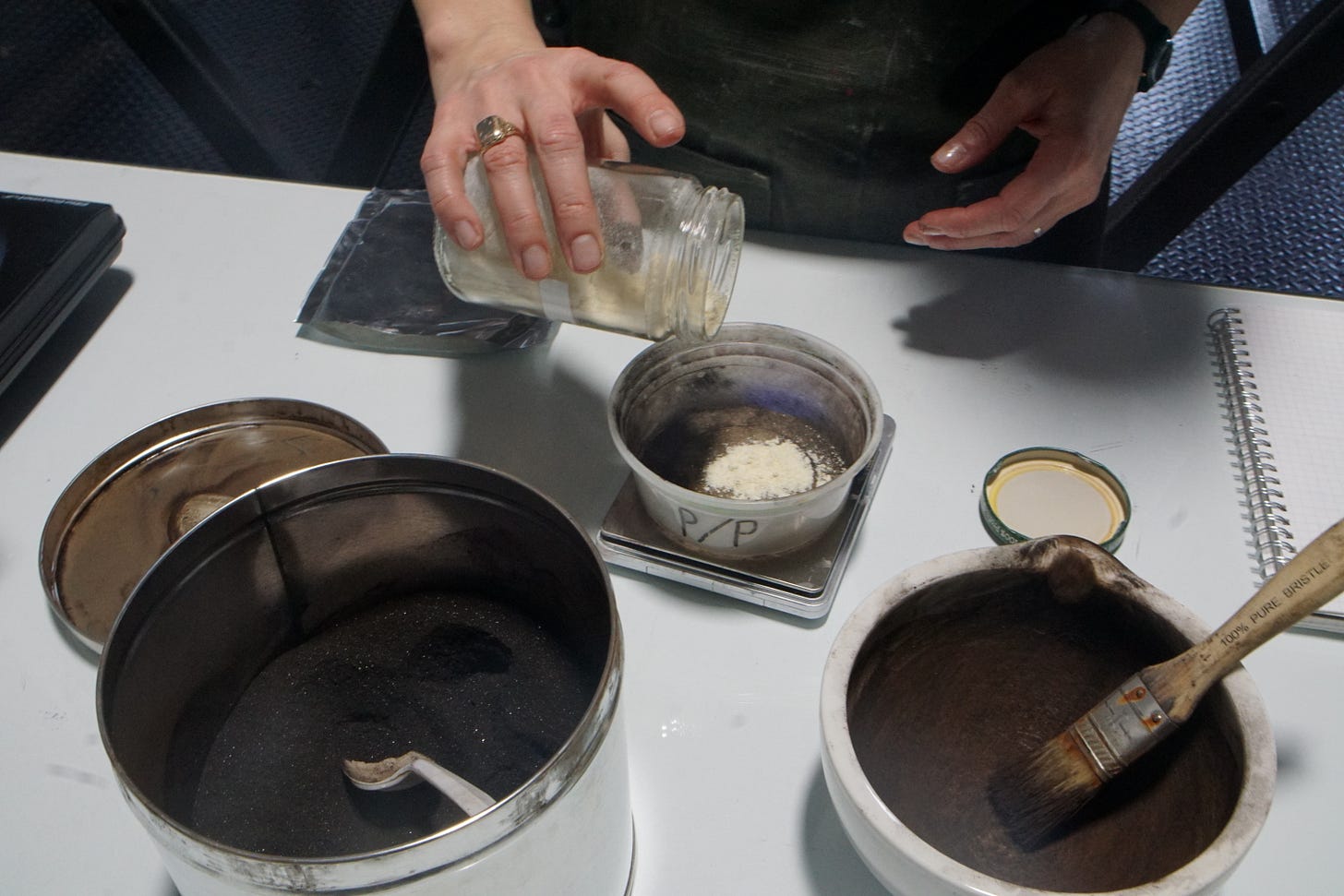 A hand holding a glass jar taps pine resin into a container on a scale. In the foreground is a large metal tin holding a dark brown powder (asphaltum) and a mortar, coated with the brown powder and holding a flat paint brush.