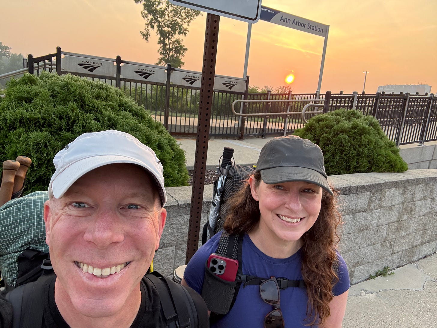 The author and her husband stand grinning in front of the Ann Arbor Station sign, about to go catch their train to Chicago and onward to Denver. The sun rises behind them amidst an eery haze caused by Ontario wildfire smoke.