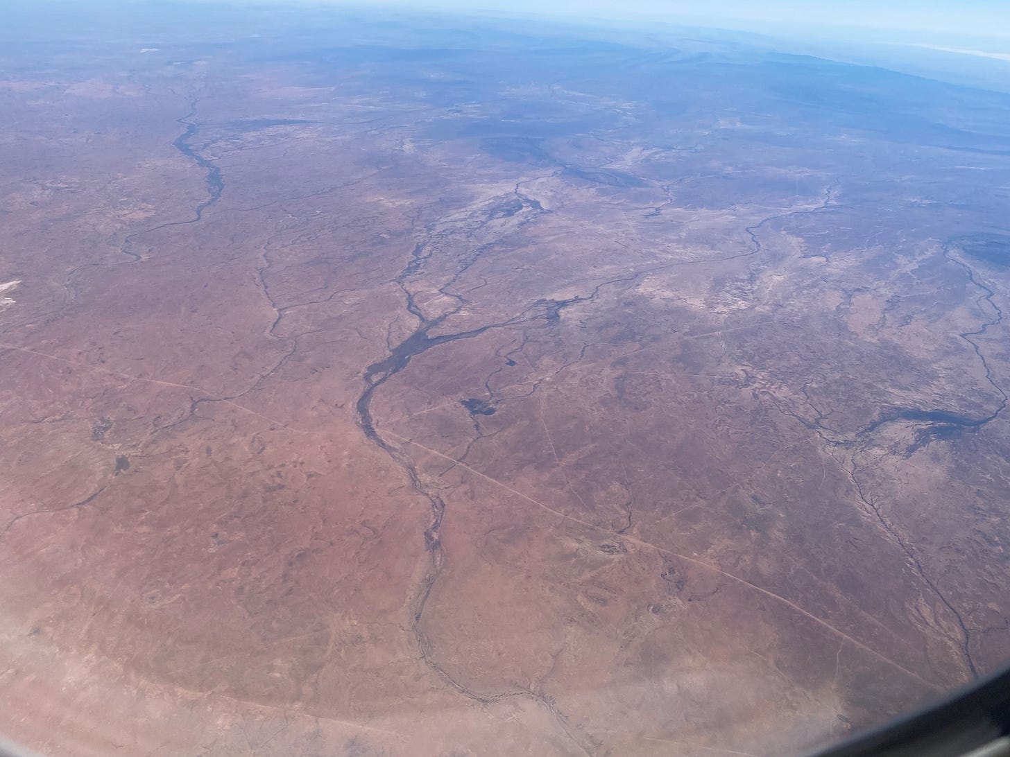 A photo out of an airplane window looking out at brown land and dark blue rivers.