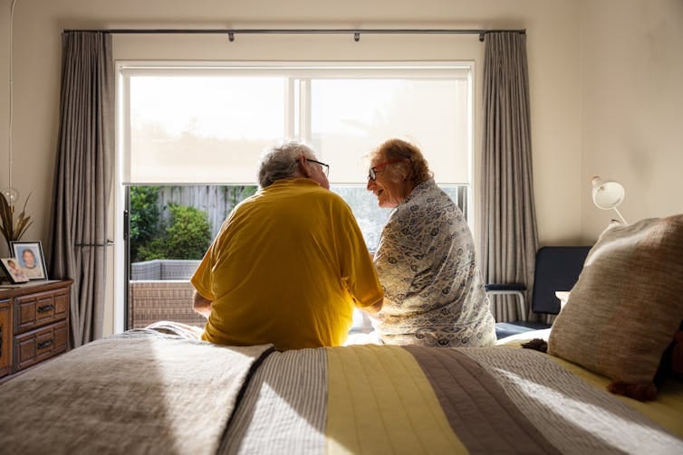 Back shot of two seniors sitting on edge of bed in front of window, speaking to one another.