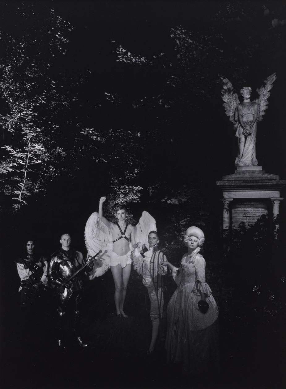 A black-and-white image shows five people in dramatic, historical costumes and a winged statue in a dark forest. The scene is mysterious and gothic. The figures from the previous images are all standing in a group on the path near the graveyard angel statue to their right. The angel has their fist in the air, and everyone else is standing confidently towards the camera, 2 on either side of the central angel.