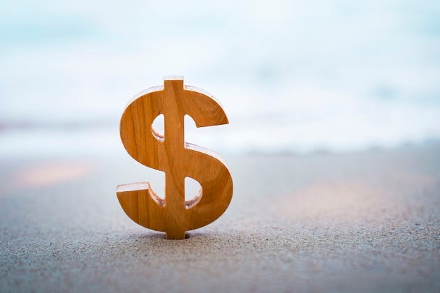Wooden dollar sign symbol standing on sand beach with sea background. Concept of financial freedom, wealth management, offshore banking, investment, and retirement plan.