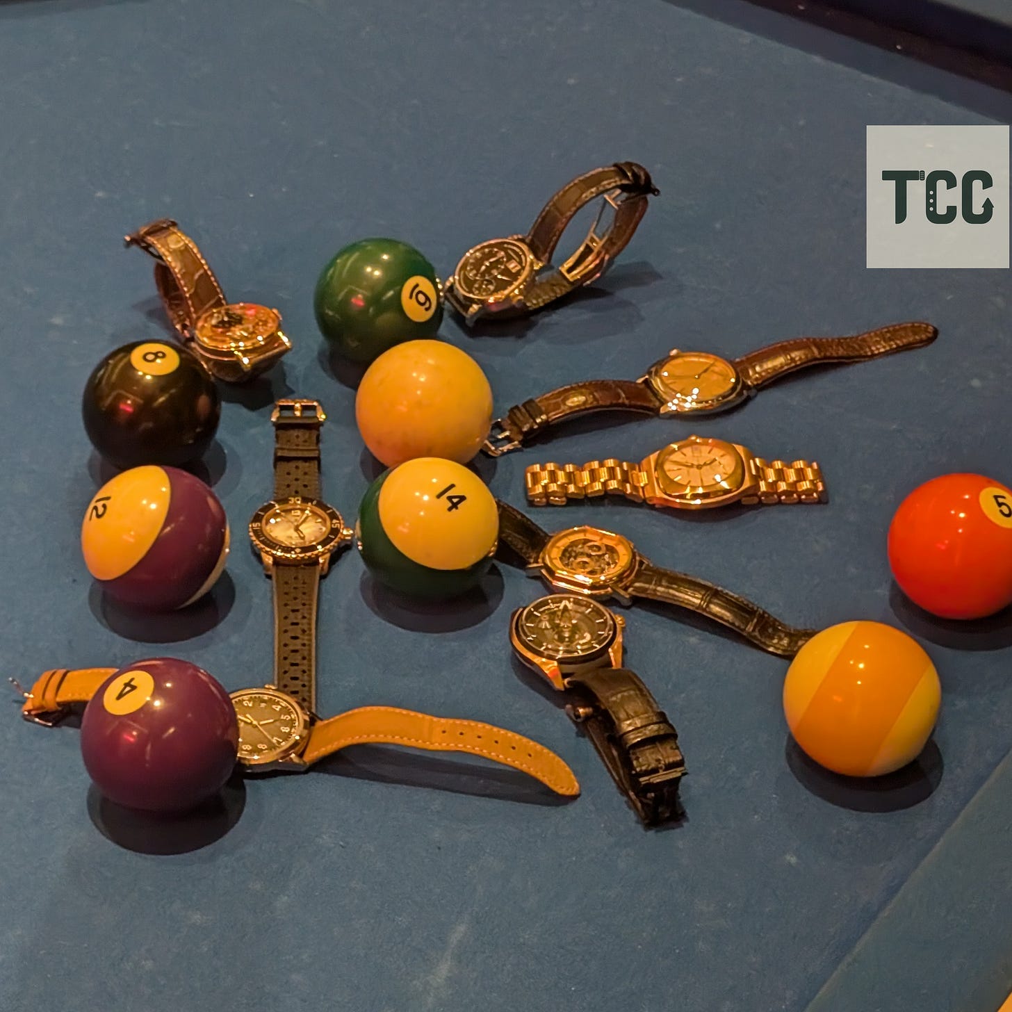 A playful scene showing a collection of wristwatches laid out among billiard balls on a blue felt pool table. The watches vary in design, including leather straps, metal bracelets, and open-worked dials. Two hands and an “INDIES.NYC” wristband are visible in the top corner of the frame, adding to the candid nature of the shot.