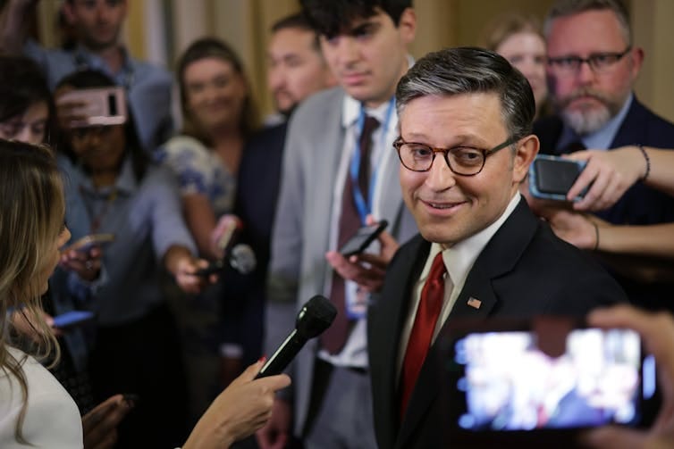 A white man with glasses, dark hair and a dark suit with a white shirt and red tie smiles and appears to speak into a microphone as people surround him. A white man with glasses, dark hair and a dark suit with a white shirt and red tie smiles and appears to speak into a microphone as people surround him.