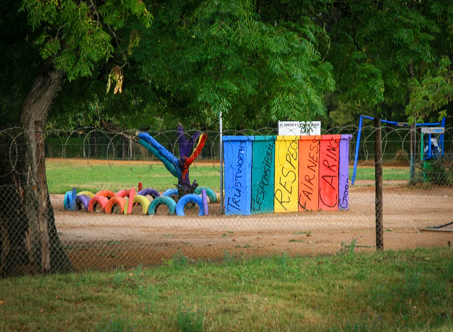 A South African schoolyard — rainbow-painted panels declaring Trustworthy, Responsibility, Respect, Fairness, Caring — seen through a fence topped with razor wire. A South African schoolyard — rainbow-painted panels declaring Trustworthy, Responsibility, Respect, Fairness, Caring — seen through a fence topped with razor wire.
