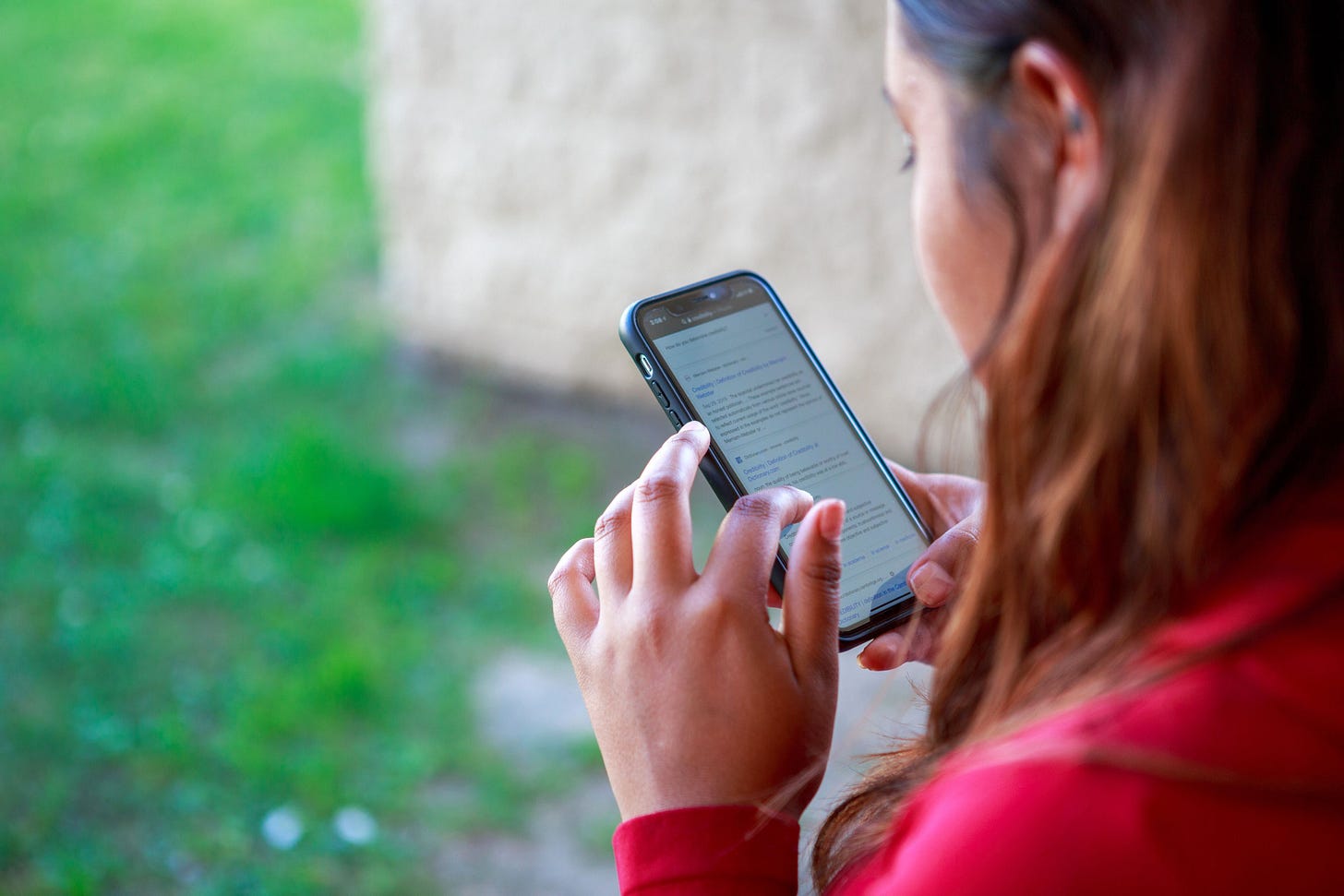 A high school student with long brown hair, wearing a red top, is holding a smartphone and looking at the screen while standing outside. The phone displays text in a web browser, and she is using her other hand to scroll. A blurred wall and green grass are visible in the background.