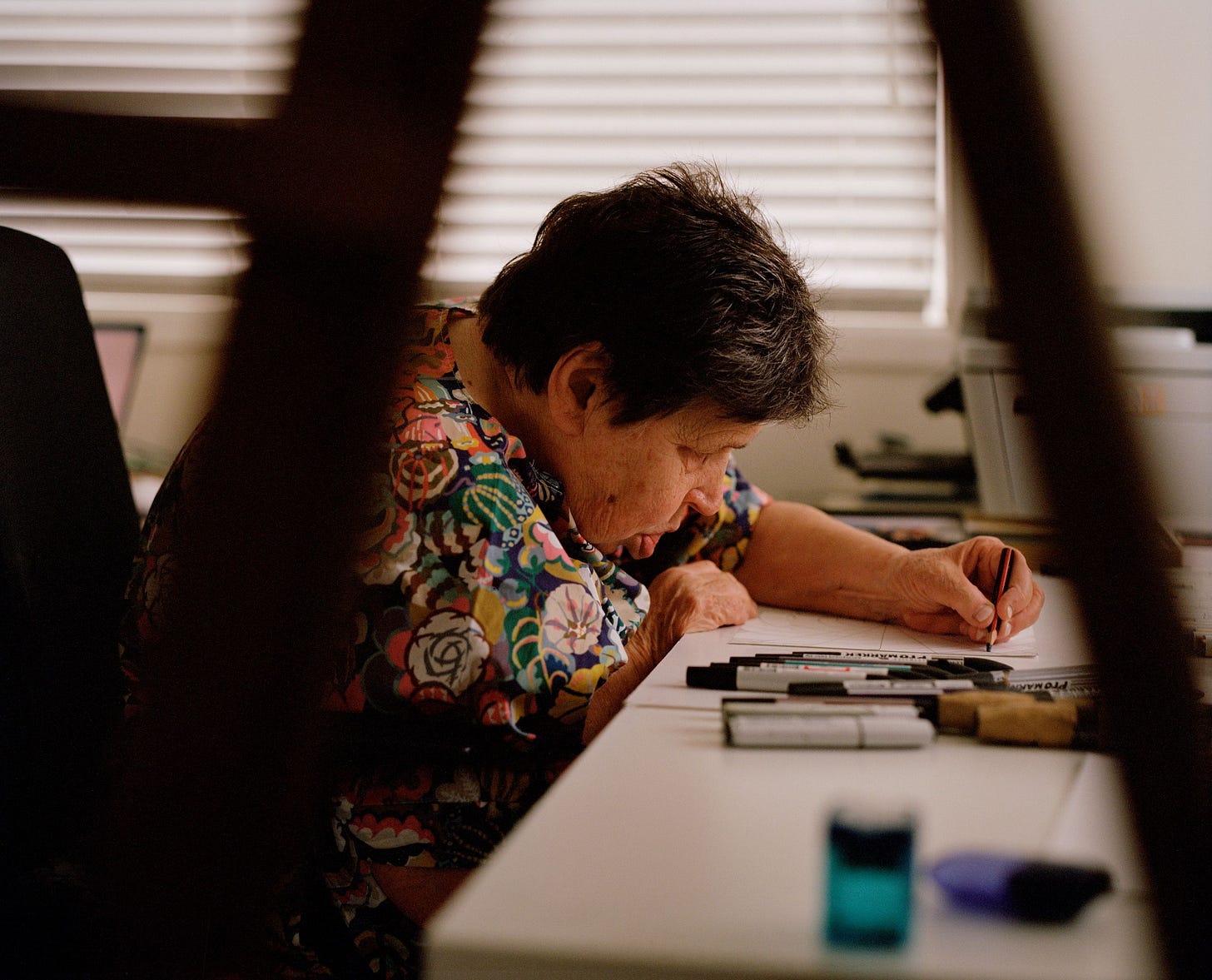 A photo of a woman in her 70s drawing at a desk.