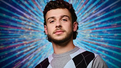 A head and shoulders shot of George Clarke, wearing a grey jumper with a blue and black diamond pattern, set against a glittering blue background