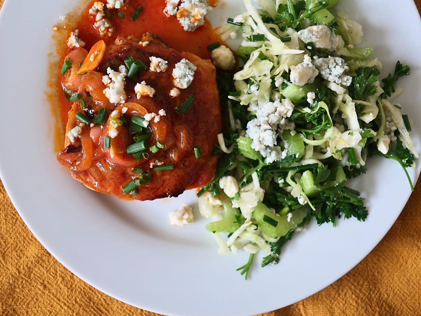 An overhead view of a Buffalo-sauce-coated chicken thigh topped with chives and blue cheese crumbles on a plate with a portion of shredded cabbage salad studded with celery, herbs, and more blue cheese crumbles.