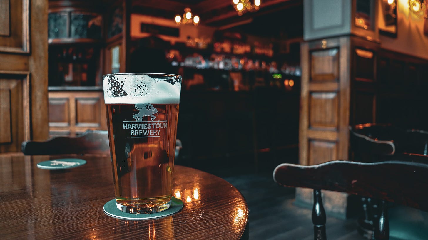 A pint of bitter on a wooden table with an ornate pub in the background.