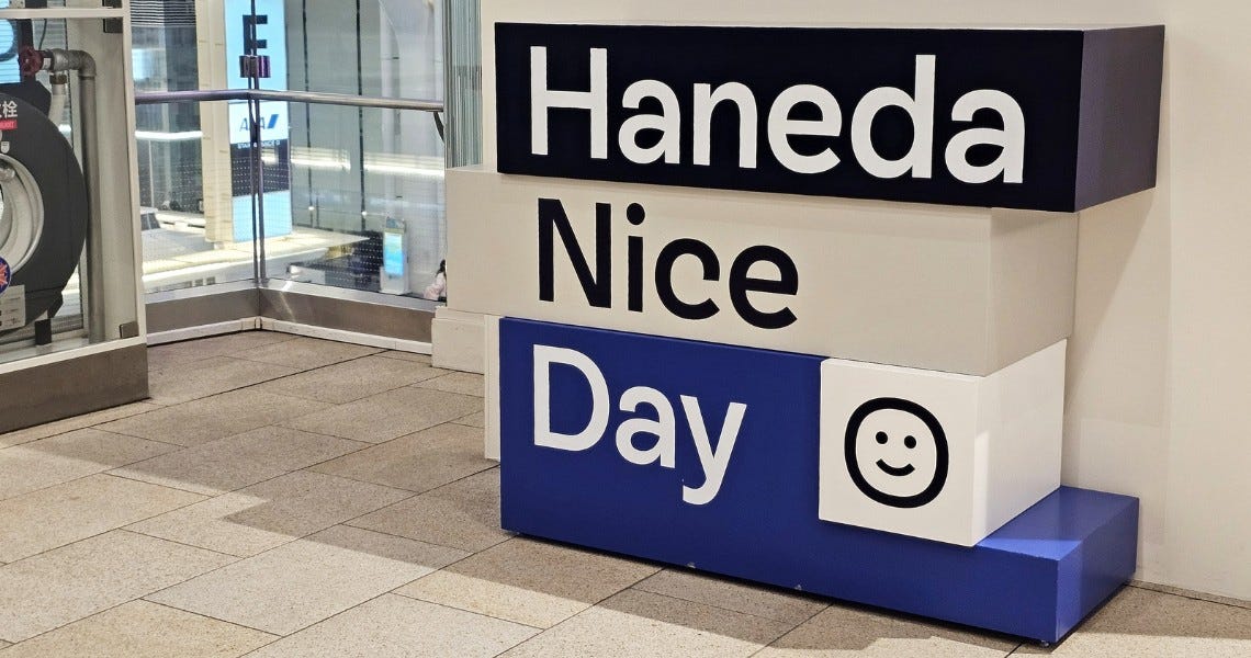 **A decorative sign installation at Haneda Airport in Tokyo, Japan.** The display consists of three stacked rectangular blocks in navy blue and white, reading "Haneda Nice Day" with a simple smiley face icon on the bottom right block. The sign is positioned in what appears to be a terminal corridor, with glass doors and tiled flooring visible in the background.